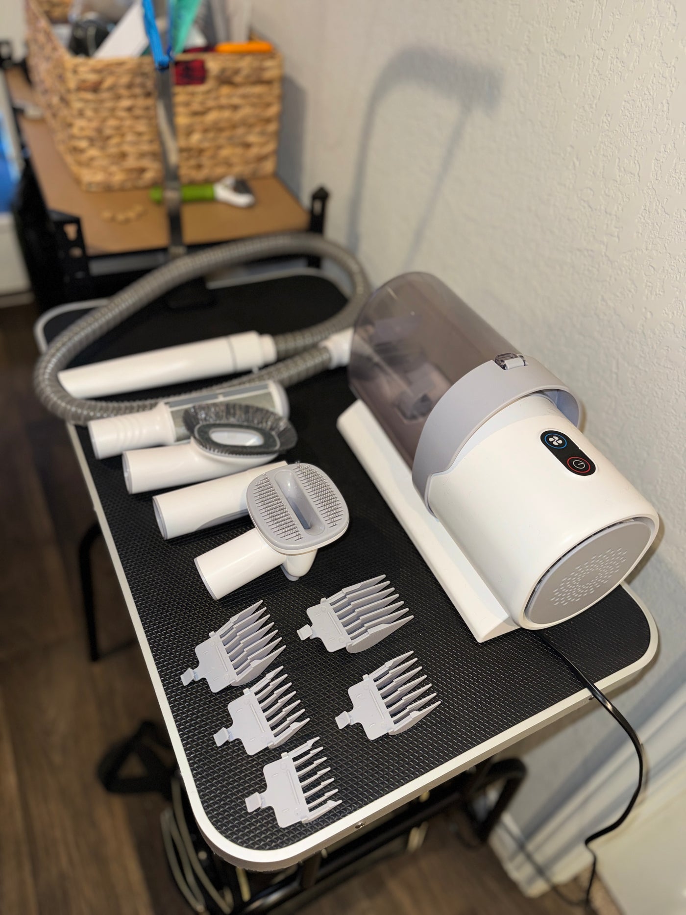 A grooming chair attachment kit with a white and gray electric grooming device, a clear hose, several white attachment heads, and multiple white comb guards arranged on a black textured grooming table.