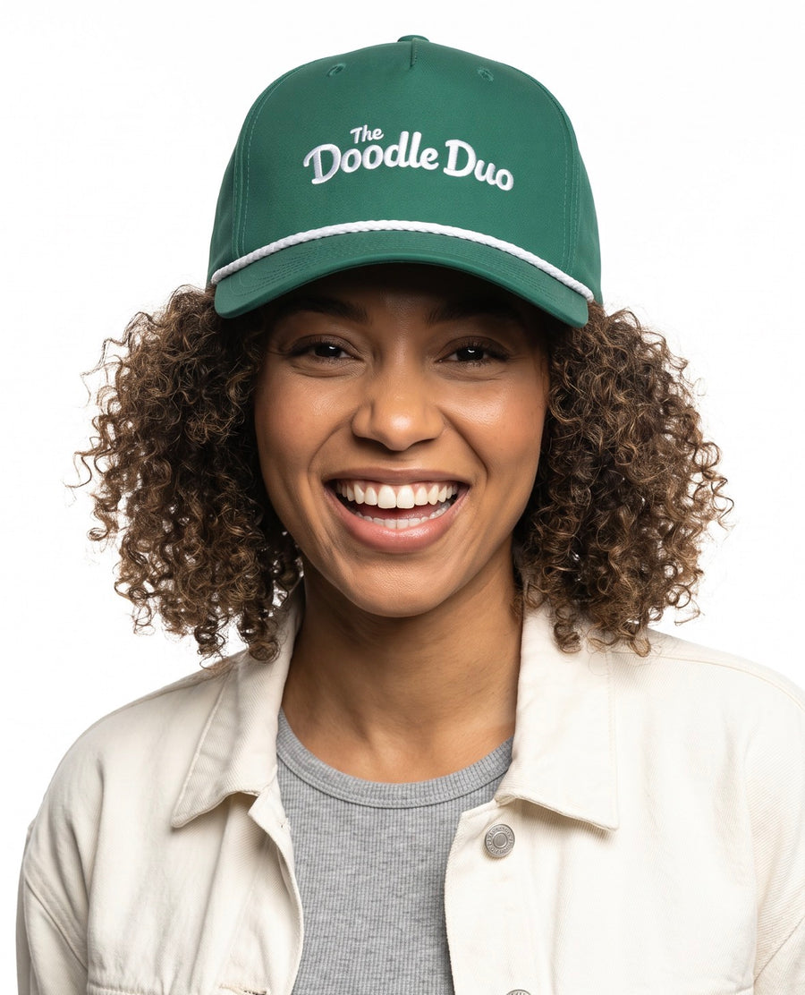 A smiling woman with curly brown hair wearing a green baseball cap with white embroidered text reading 'The Doodle Duo' and a white stripe detail on the brim, paired with a cream jacket over a gray shirt, against a plain white background.