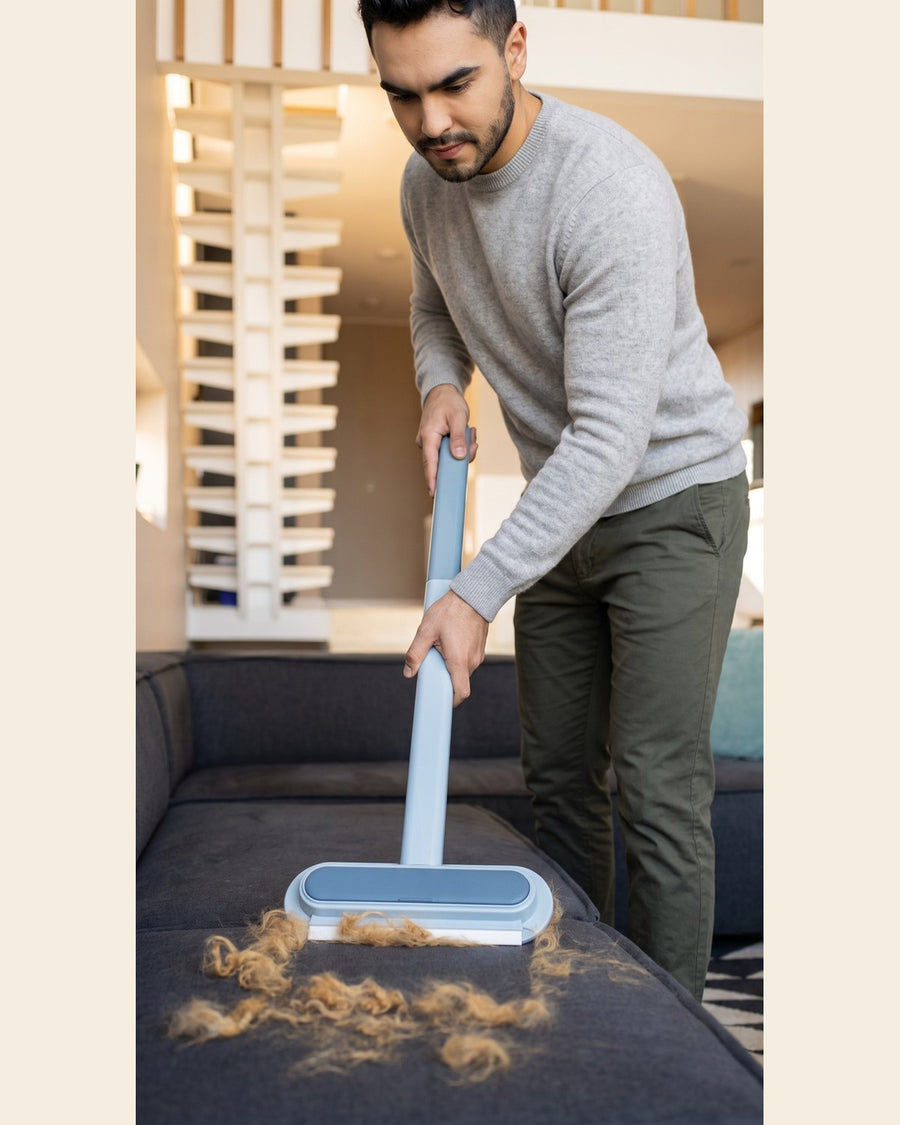 A man in a gray sweater and dark pants uses a light blue pet hair remover tool on a dark sofa, clearing out a pile of tan fur.