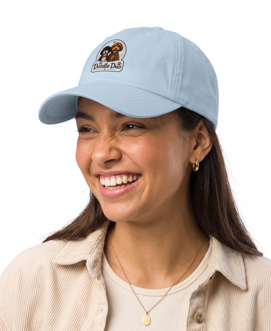 A smiling woman wearing a light blue baseball cap with an embroidered 'Doodle Dog' logo, paired with a beige corduroy shirt against a plain white background.