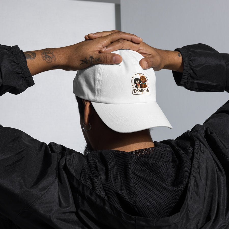 A person wearing the white Paw's Hat dad cap with The Doodle Duo logo patch on the front, photographed from behind in a black windbreaker.