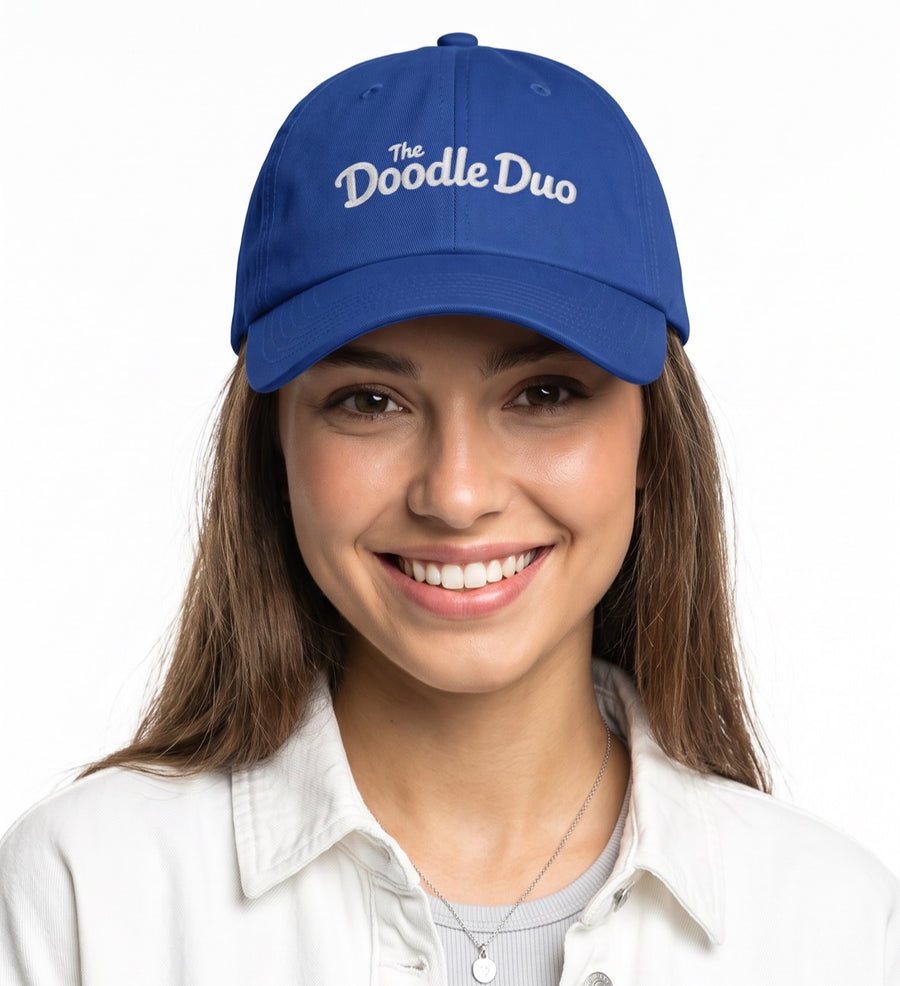 A smiling woman wearing a royal blue baseball cap with 'The Doodle Duo' embroidered in white script on the front, paired with a white button-up shirt against a plain white background.