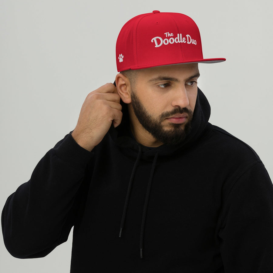 The Doodle Duo red snapback hat with white signature wordmark and paw print embroidery worn by a man in a hoodie.