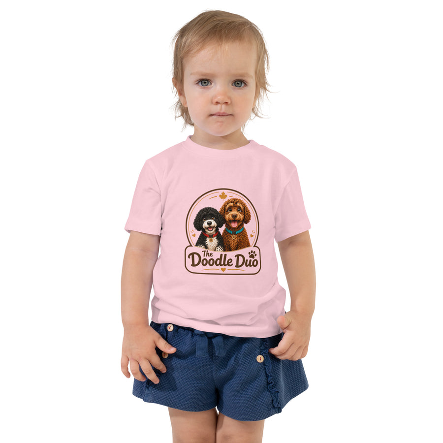 A toddler standing against a white background wearing a light pink short-sleeve t-shirt with a 'The Doodle Duo' graphic print on the front, paired with navy blue shorts.